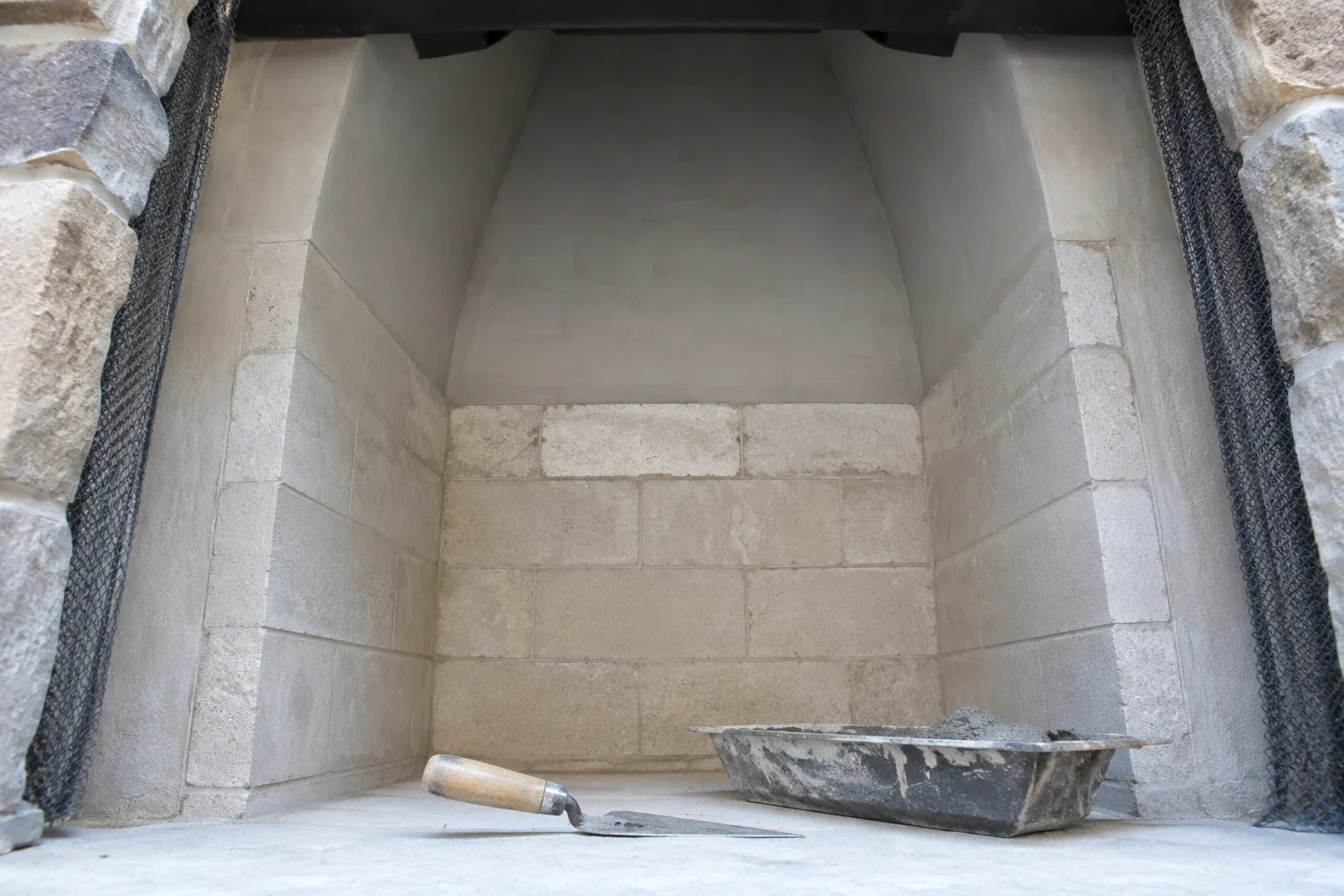 Pale masonry fireplace interior with sloping upper walls, a trowel and a tray of gray material on the hearth.